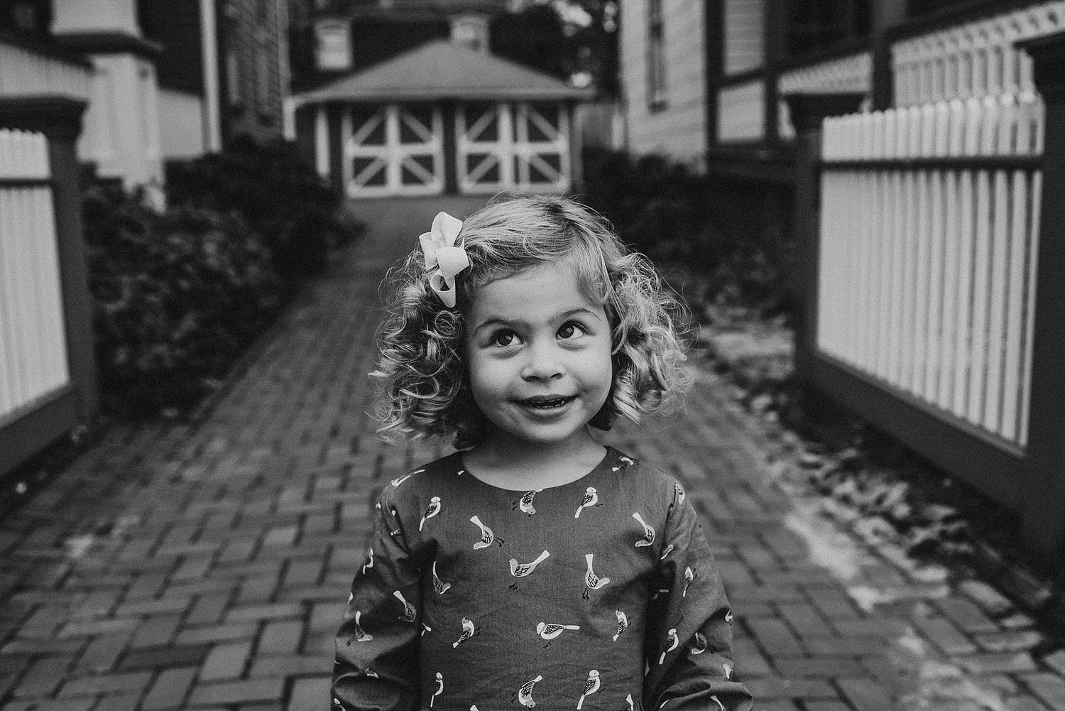 A black and white portrait of a child with curly hair on a brick pathway.
