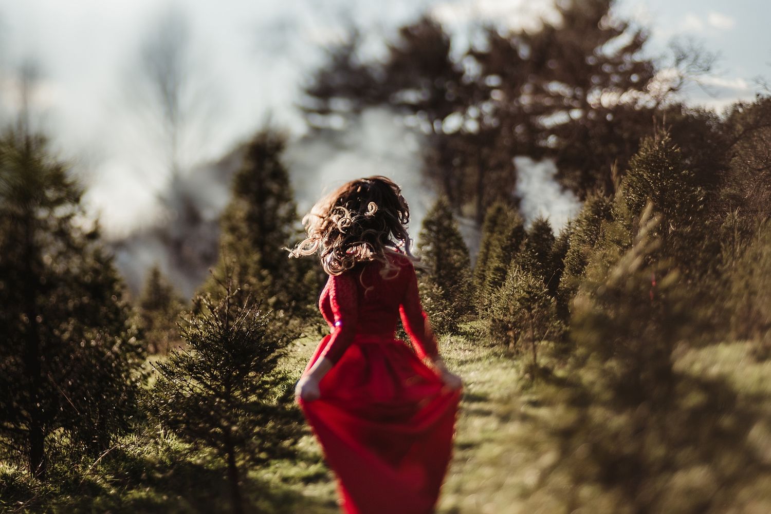 Woman in flowing red dress walking through sunlit forest path with hair blowing in the wind.