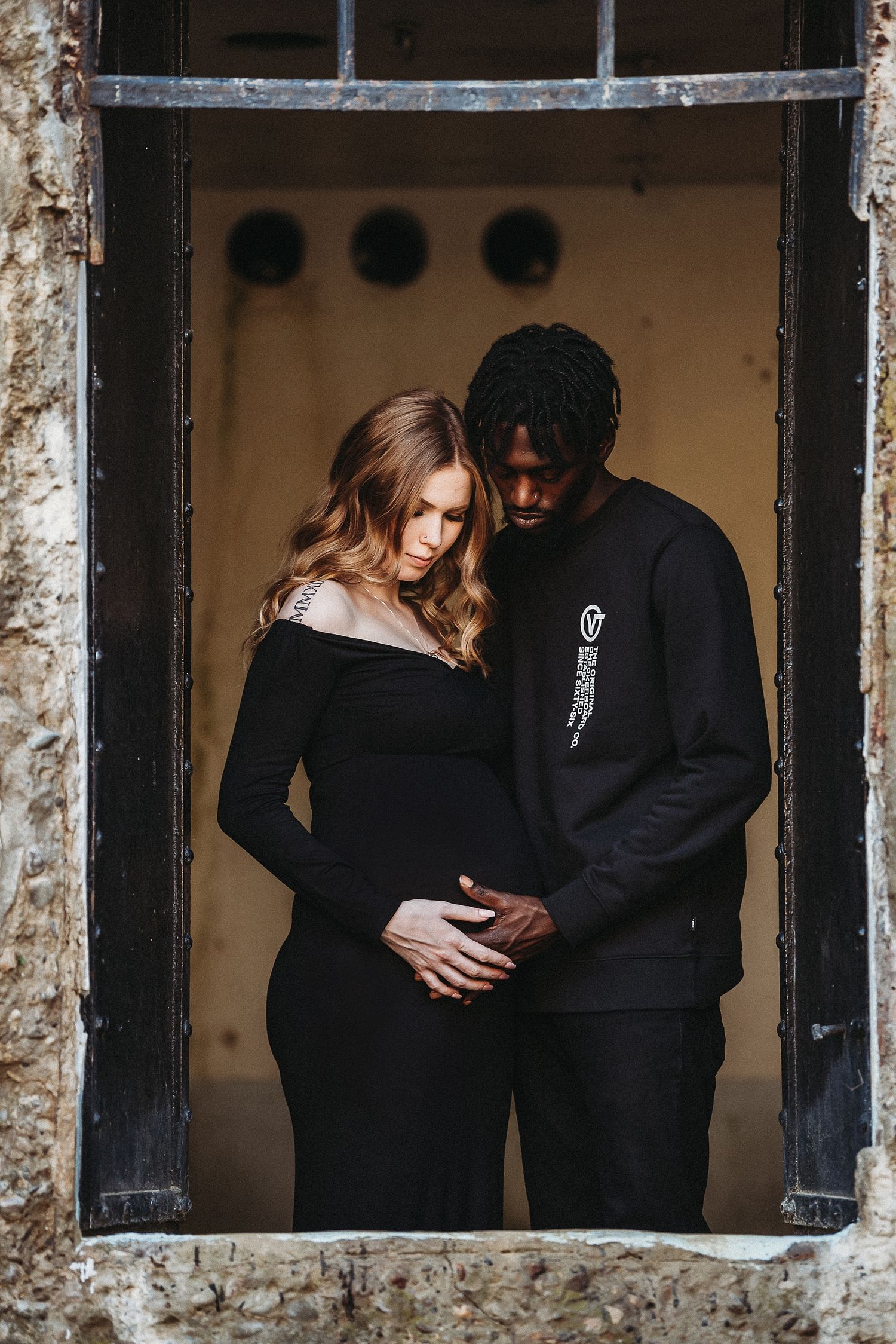 Intimate maternity photo of couple standing in window frame wearing black clothing.