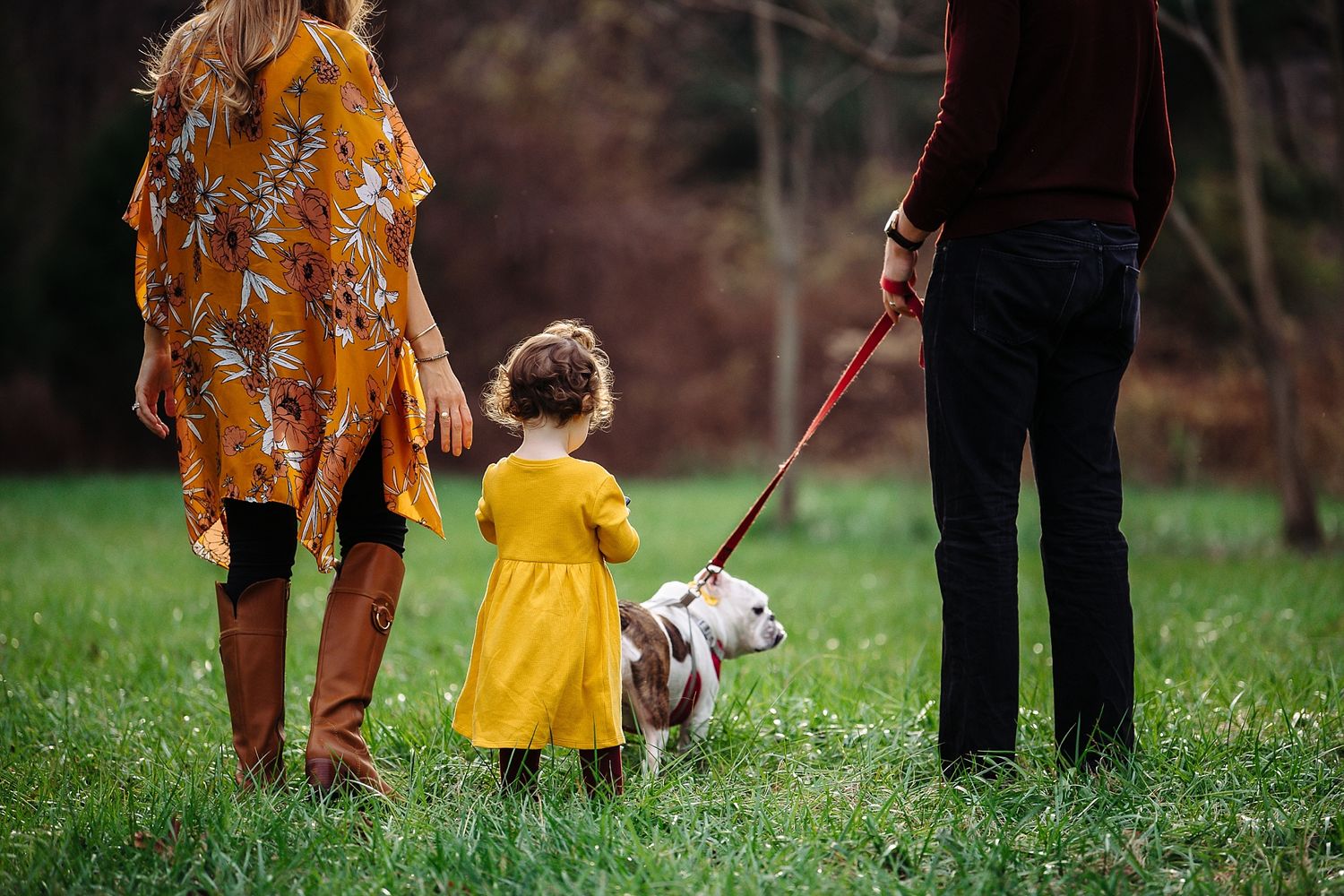 A family walks their small dog together through a grassy field on an autumn day.
