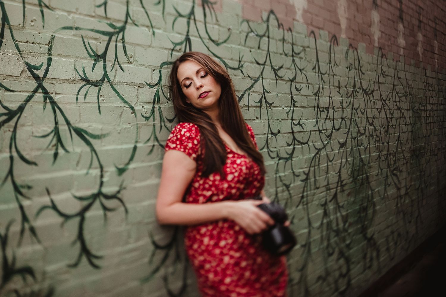 Woman in red polka dot dress with camera against urban wall with painted vines.