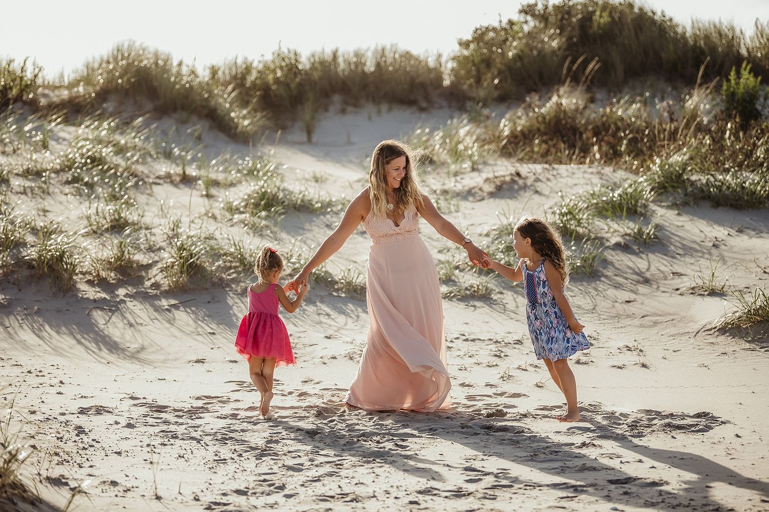 Three people hold hands while dancing together on sandy dunes at sunset.