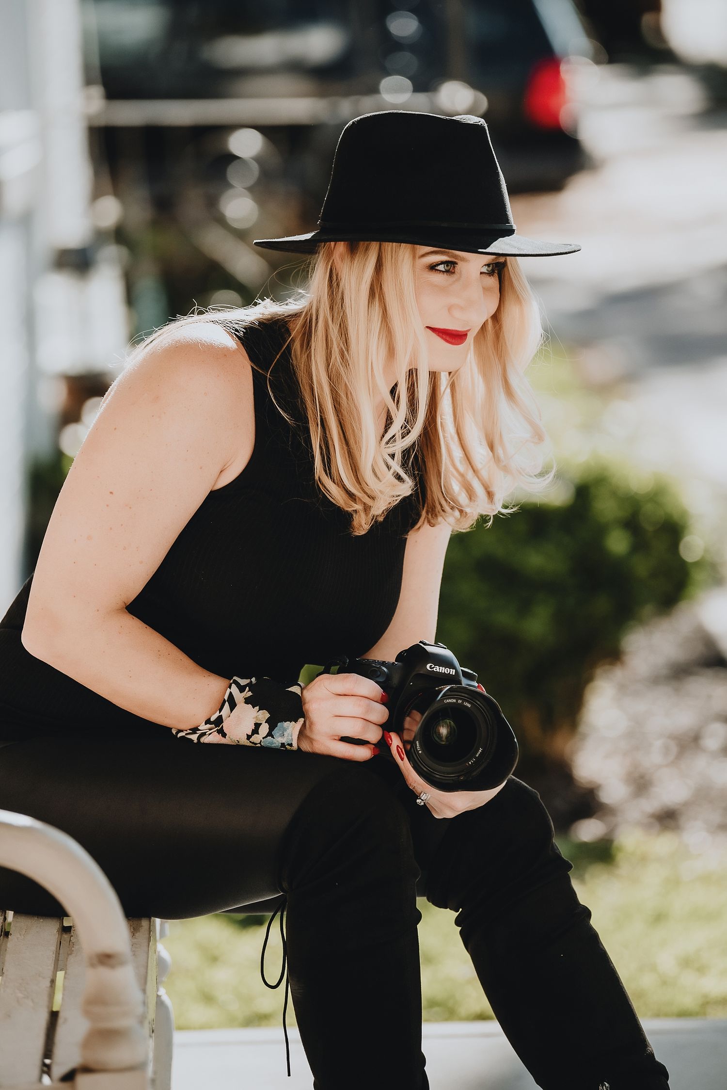 Photographer in black outfit and hat holds camera while sitting outdoors.