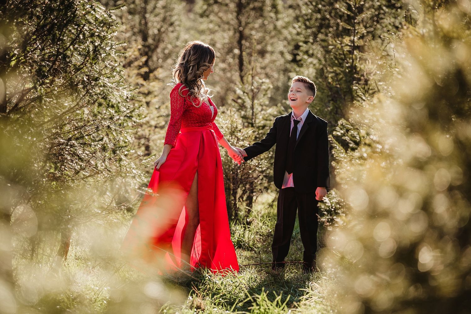 A couple shares a romantic moment in a forest, with her red dress flowing in the breeze.