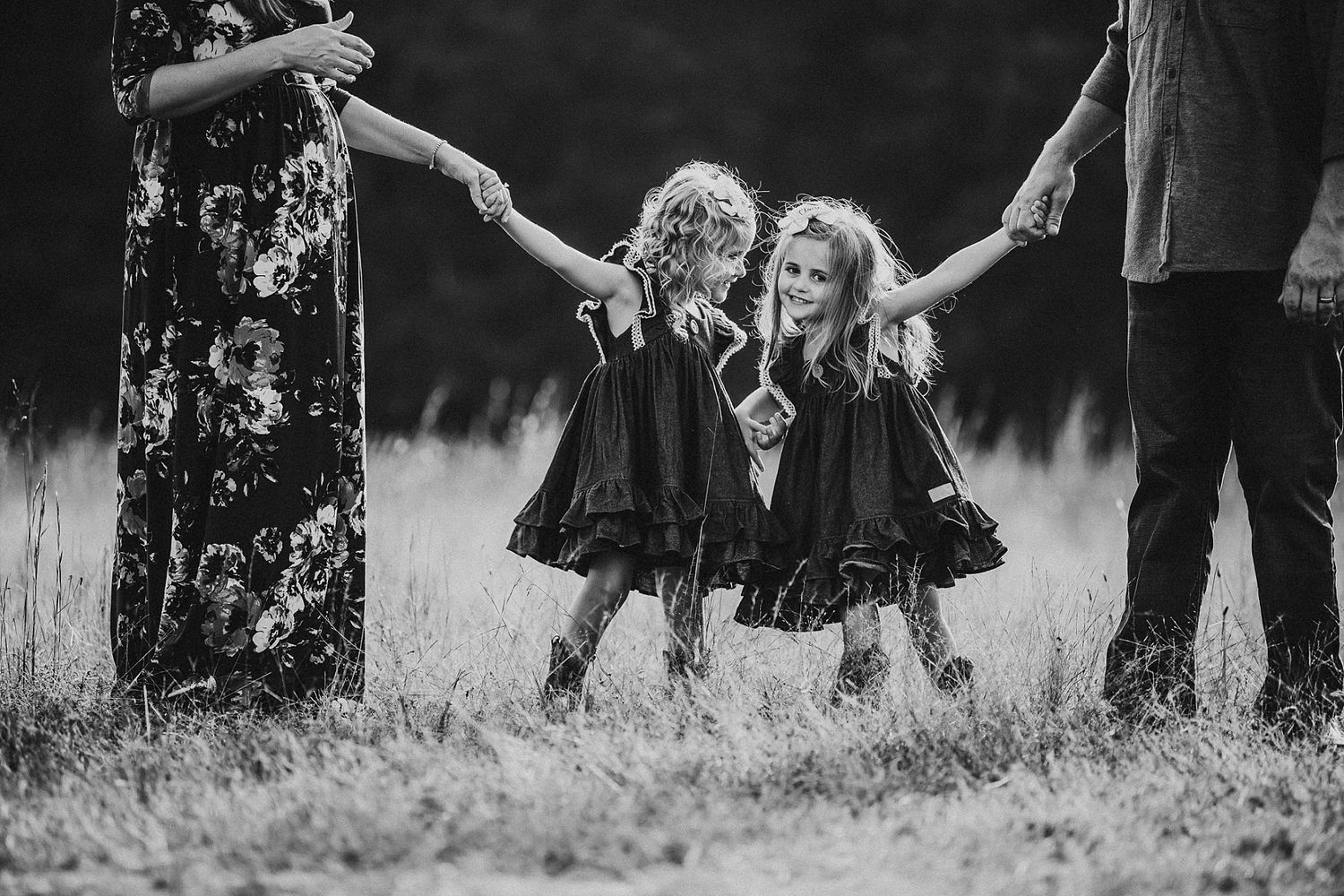 Two children in matching black dresses swing joyfully between adults in a black and white outdoor photo.