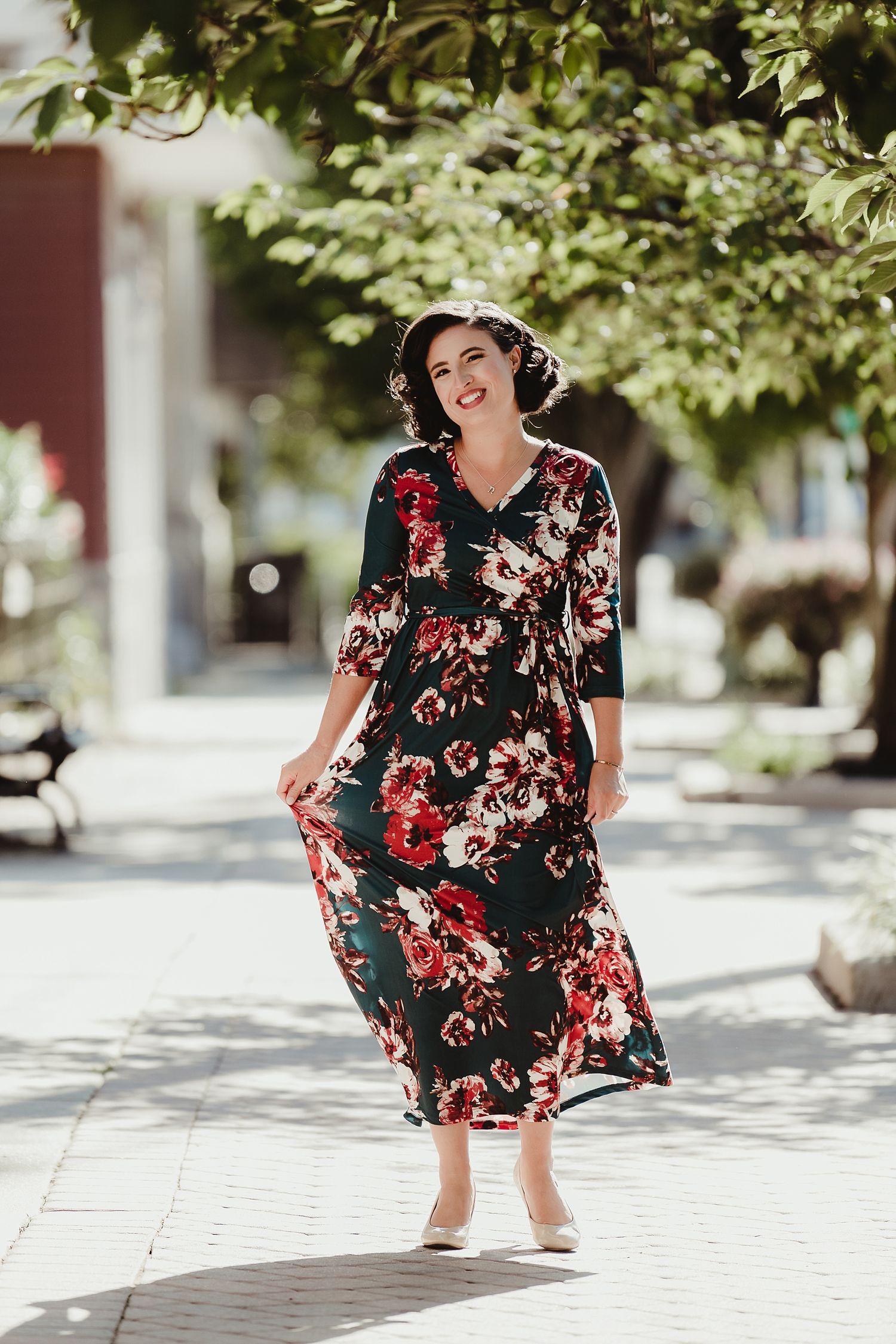 Woman in floral maxi dress twirls on sunny sidewalk with joyful expression.