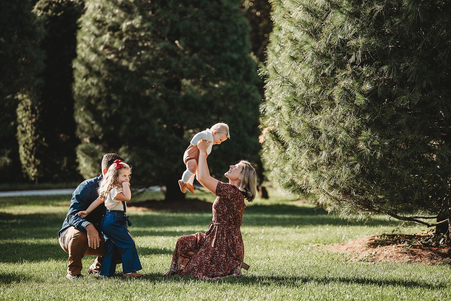 Family playing and lifting children in air on sunny lawn with evergreen trees.