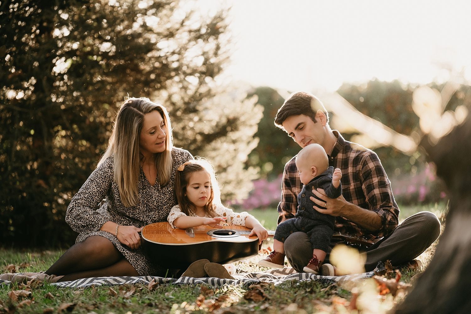 Family sitting on blanket playing guitar together during golden hour outdoor picnic.
