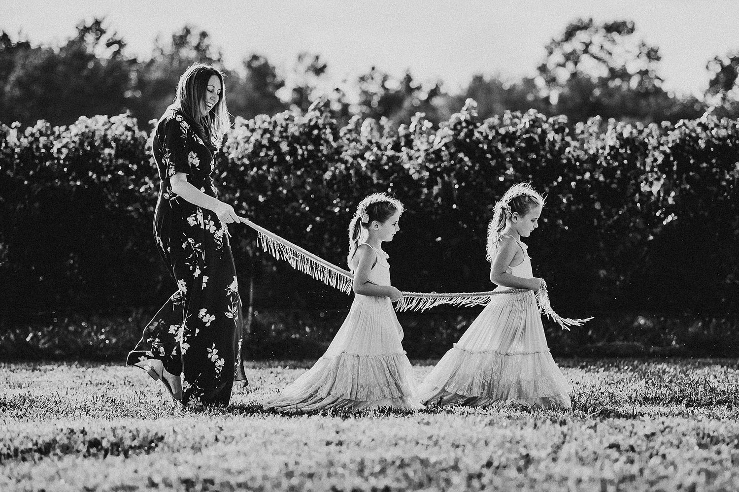 Black and white photo of parent walking with two children in matching white dresses through field.