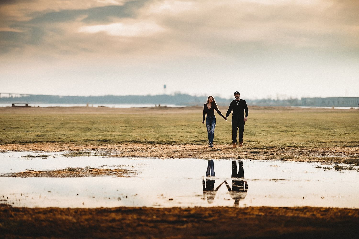 Couple holding hands reflected in puddle on cloudy beach with distant horizon.