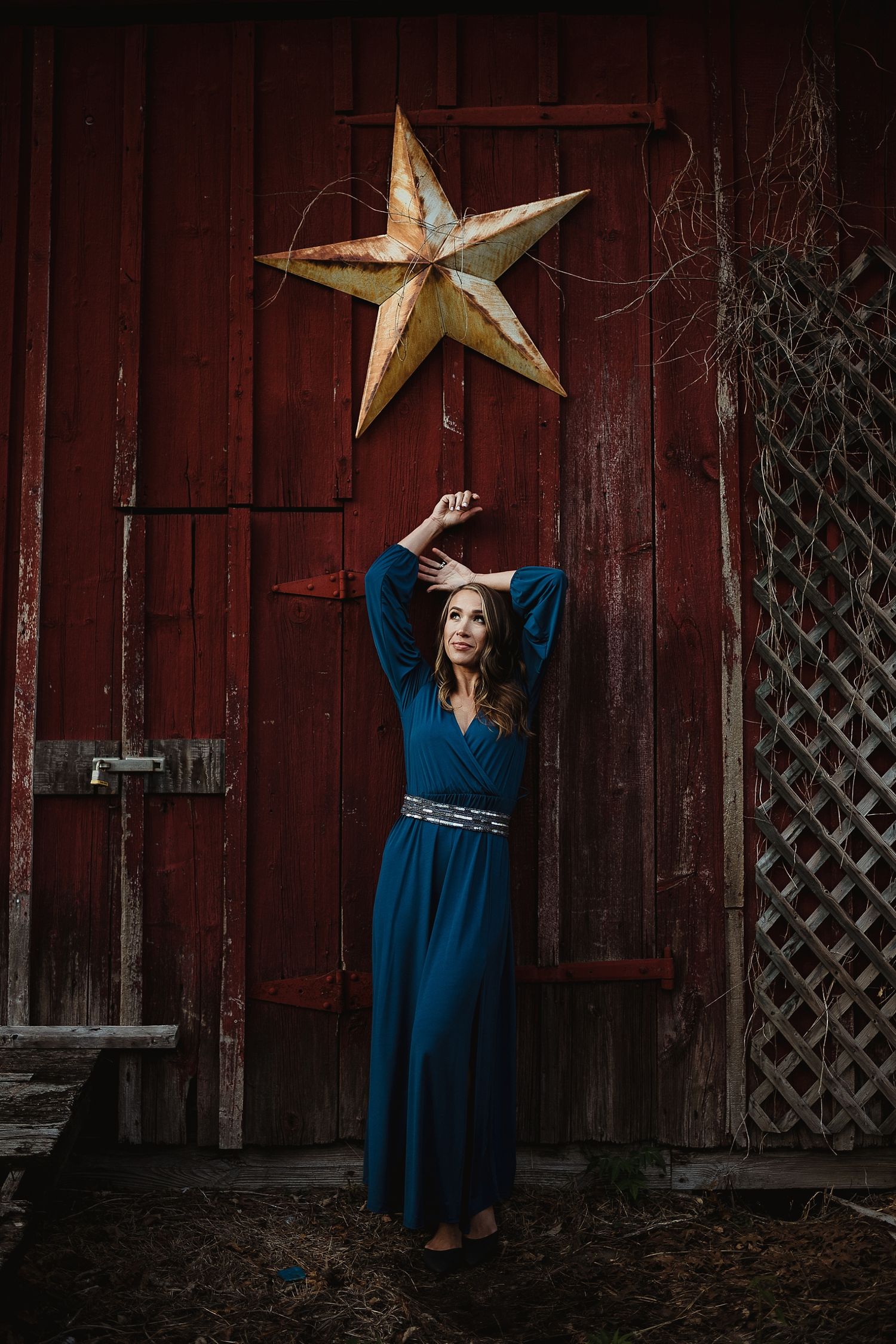 Woman in flowing blue dress poses against red barn wall with decorative metal star.
