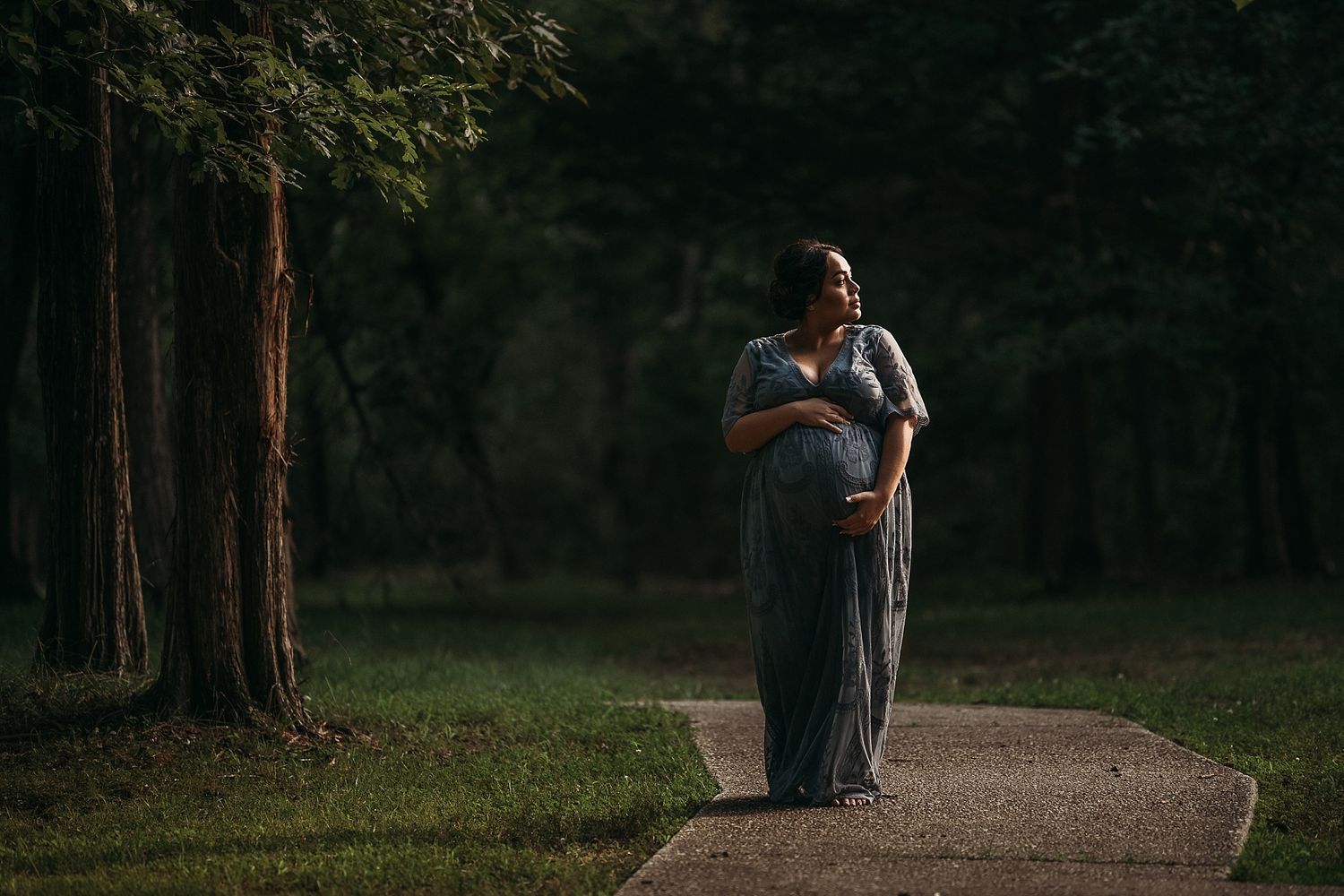 Silhouetted figure in long dress standing on path in wooded area at dusk.