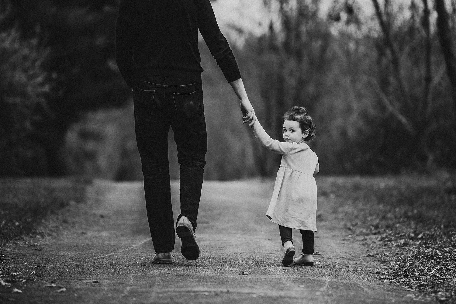 Black and white photo of a small child in a dress holding hands with an adult while walking on a woodland path.