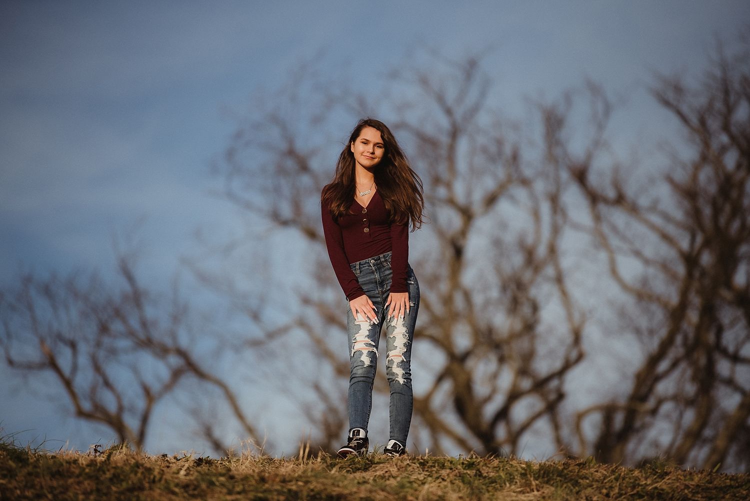 Person in ripped jeans and burgundy sweater standing on hill against winter sky with bare trees.