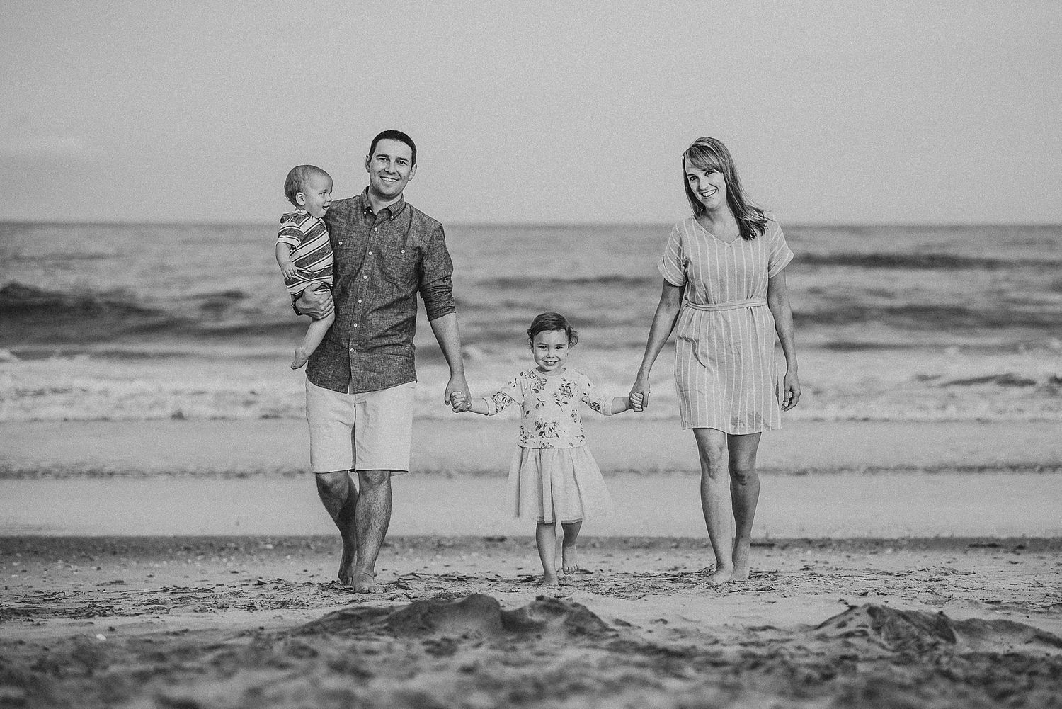 Black and white family portrait of a group standing together on a beach at sunset.