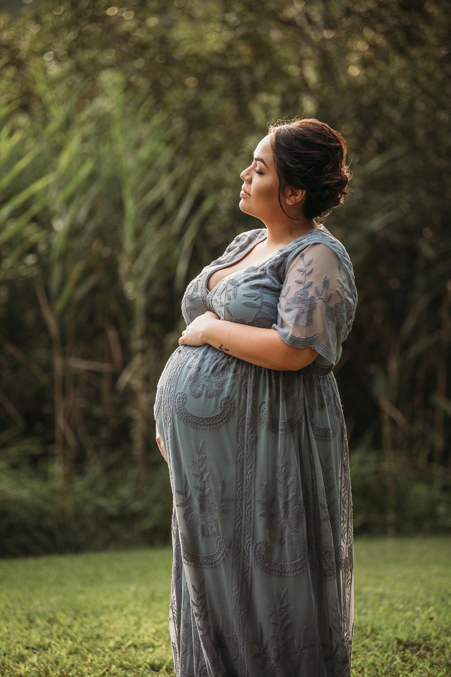 Side profile of expectant mother in flowing gray dress standing in garden during golden hour.