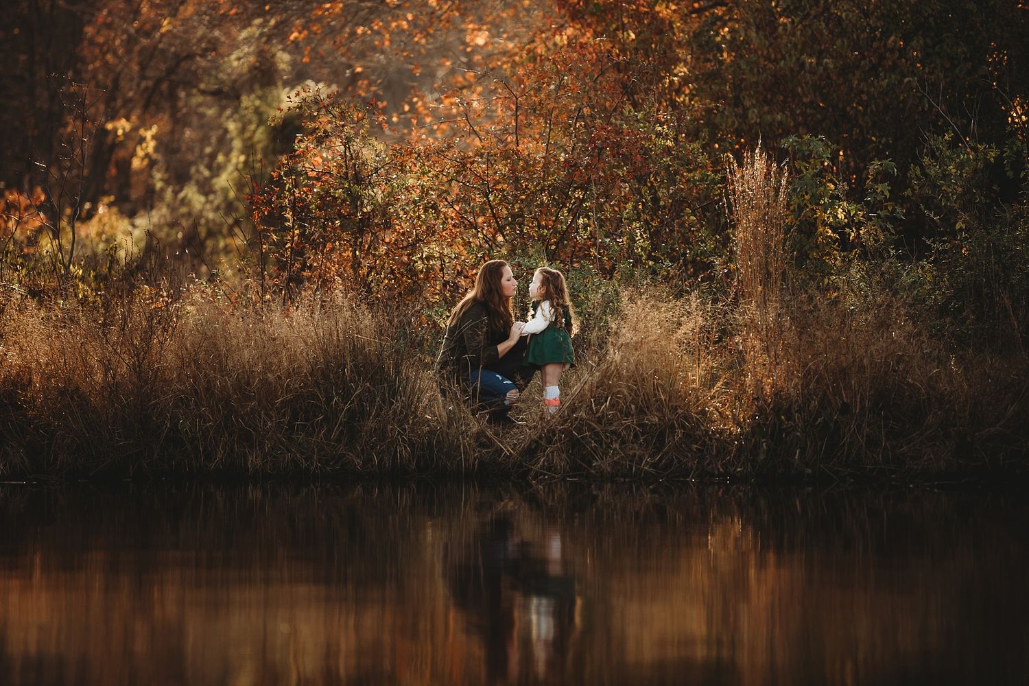 A couple sits by a tranquil pond during golden hour with autumn foliage reflected in the calm water.