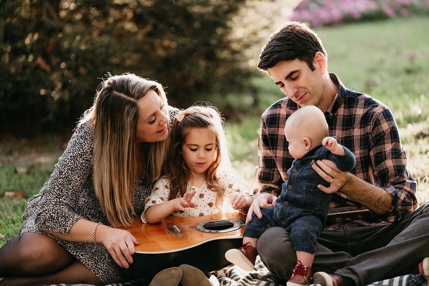 A small family sits on a blanket outdoors playing guitar together on a sunny afternoon.