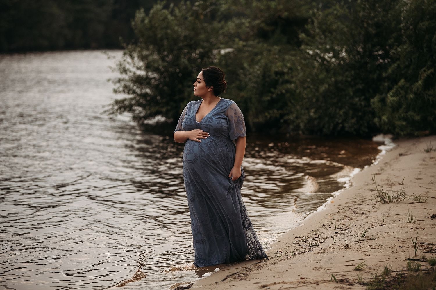 Expectant mother in a flowing blue dress stands peacefully by the water's edge during sunset.