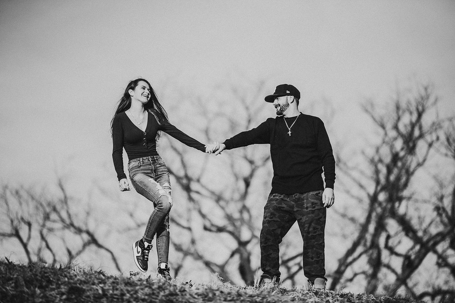 Playful couple holding hands on hilltop in dramatic black and white composition.