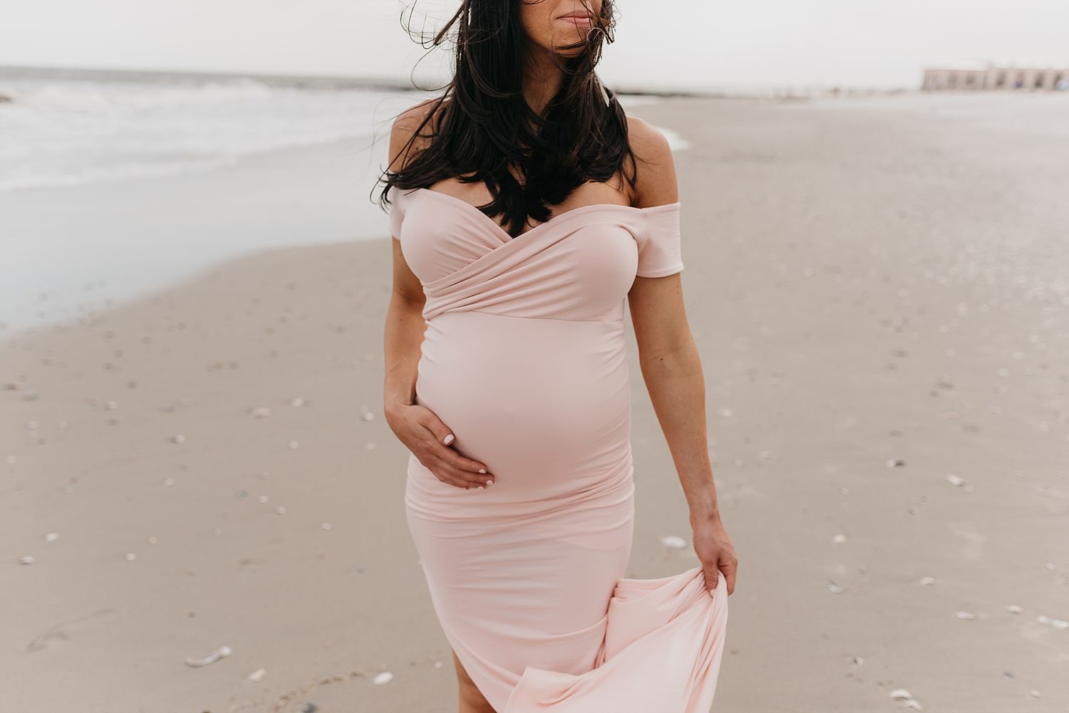 Expectant mother in pink dress walks along beach shoreline.