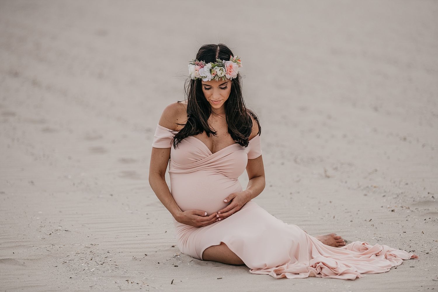 Expecting mother in flowing pink dress sits gracefully on beach sand.