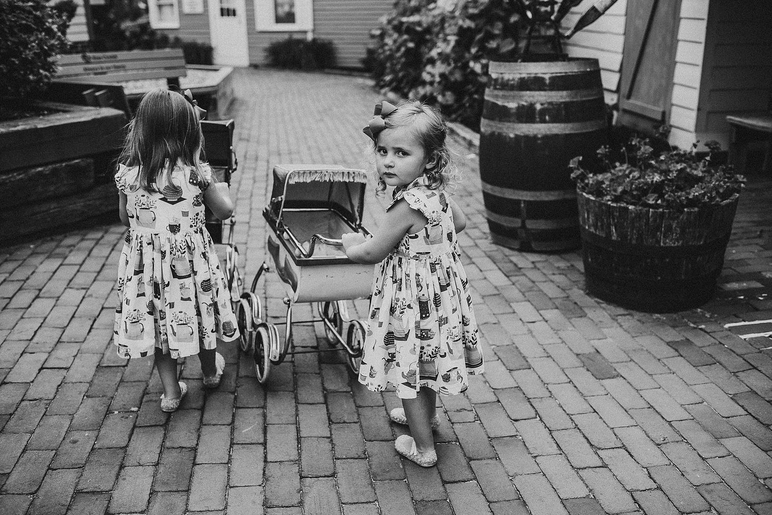 Young children in matching dresses play with vintage pram on brick patio.