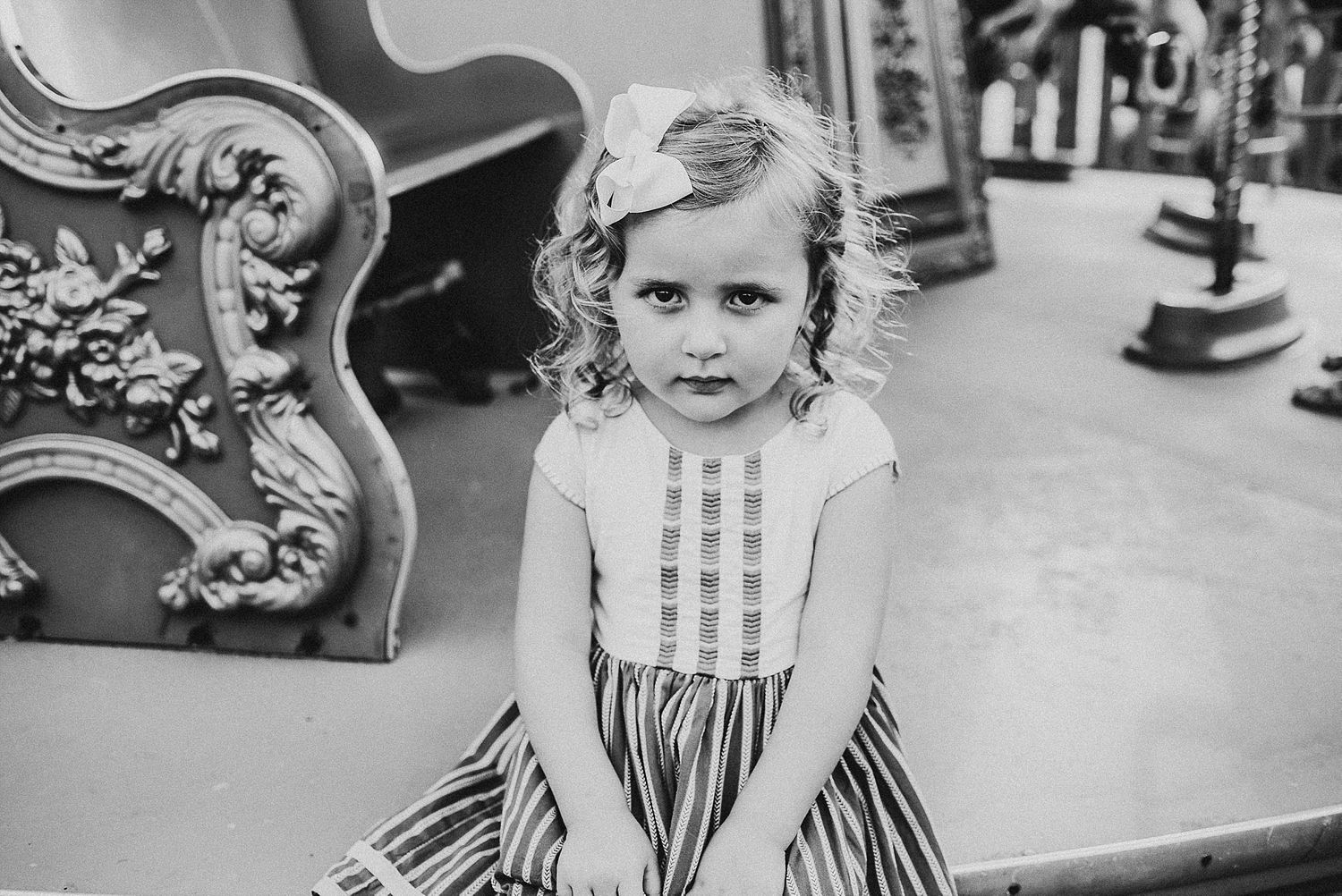 A vintage black and white photo showing a small child in a decorative dress sitting on an ornate chair.