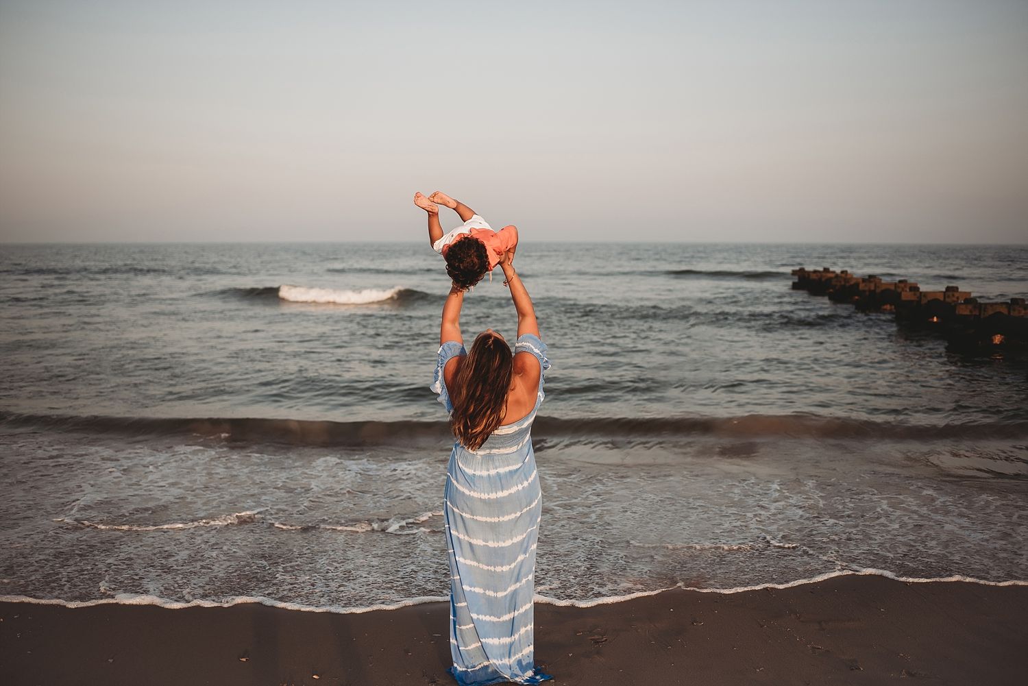 A figure in striped dress holds a child up at the beach during sunset.