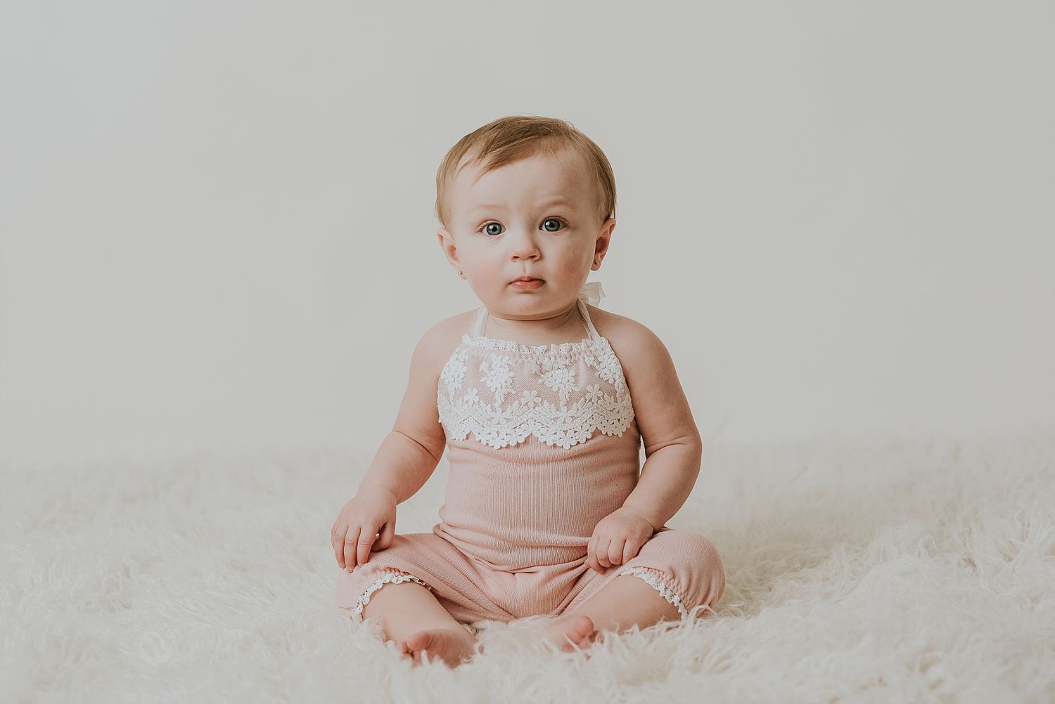 A baby in lace top and shorts sits on white fluffy rug during studio photoshoot.