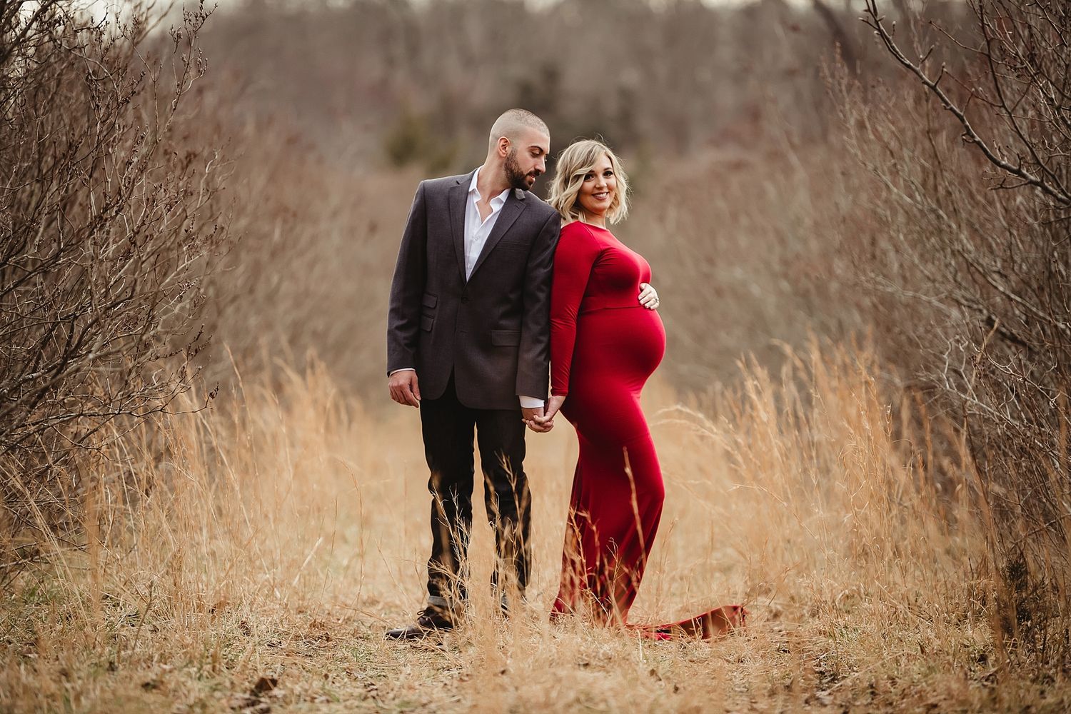A couple poses in a winter field with the woman wearing a striking red dress.
