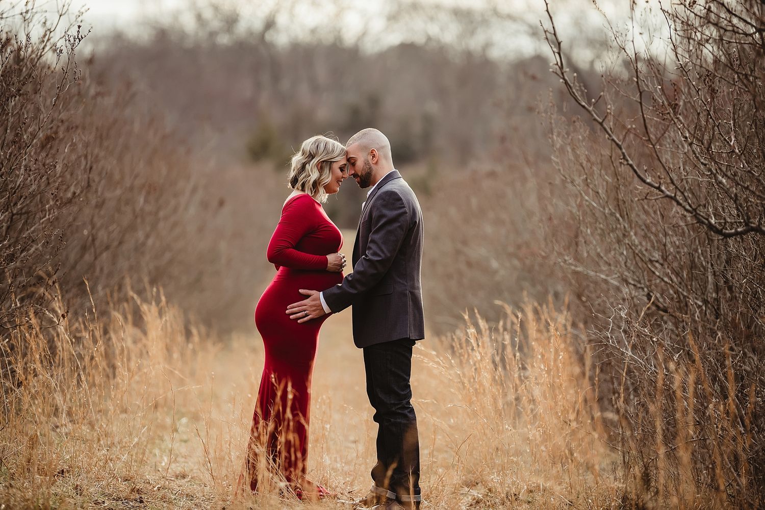A couple shares an intimate moment on a path surrounded by tall brown grass and bare branches.