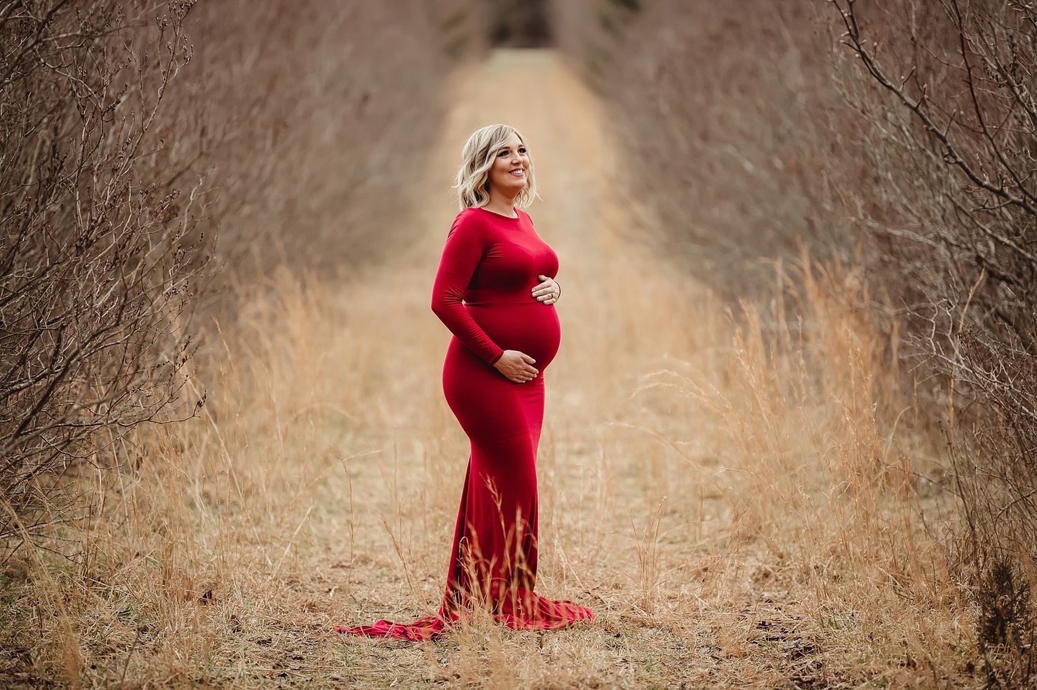 A woman in a flowing red dress poses on a path surrounded by dried brush at sunset.
