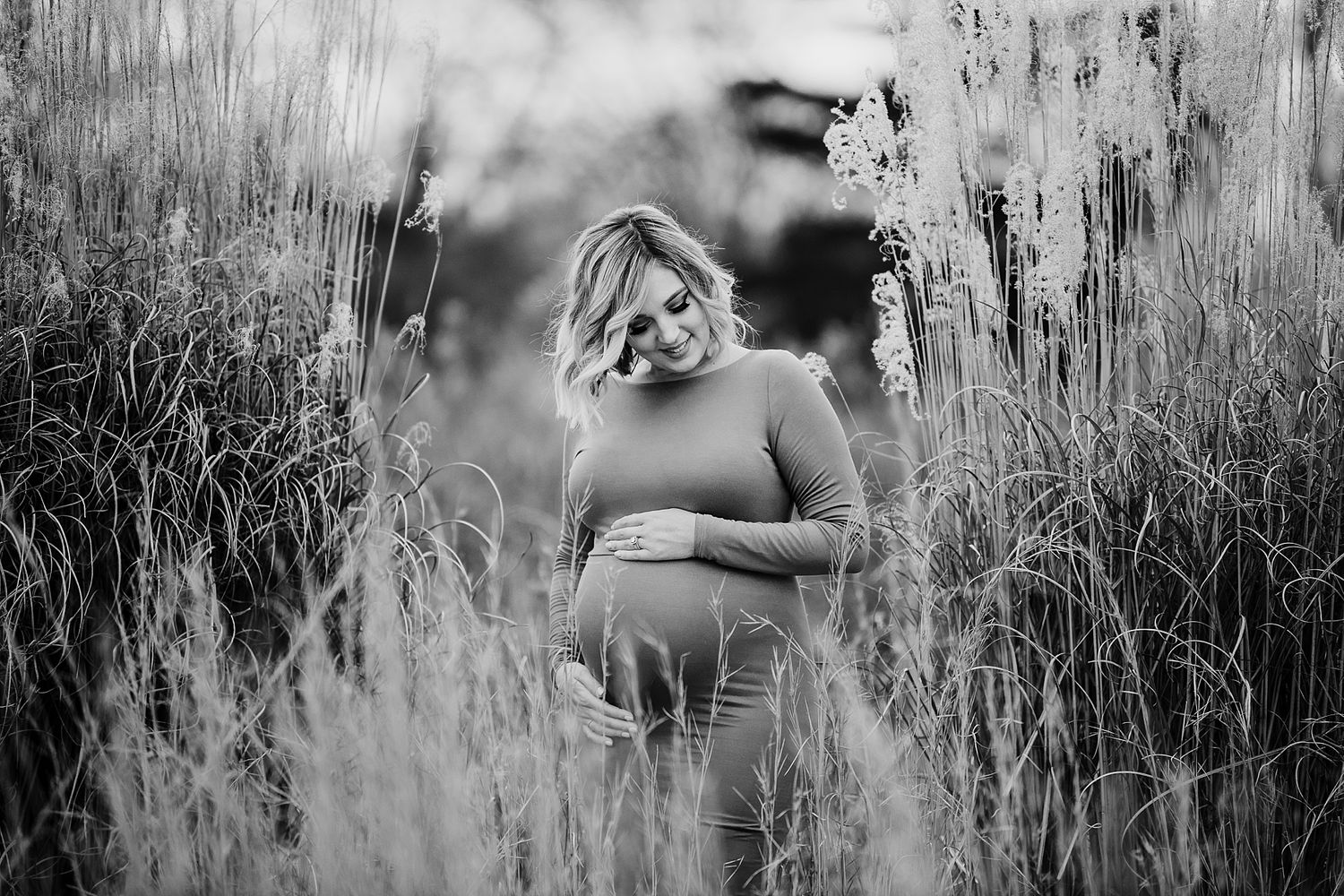 Artistic black and white maternity photo of an expectant mother standing in tall grass.