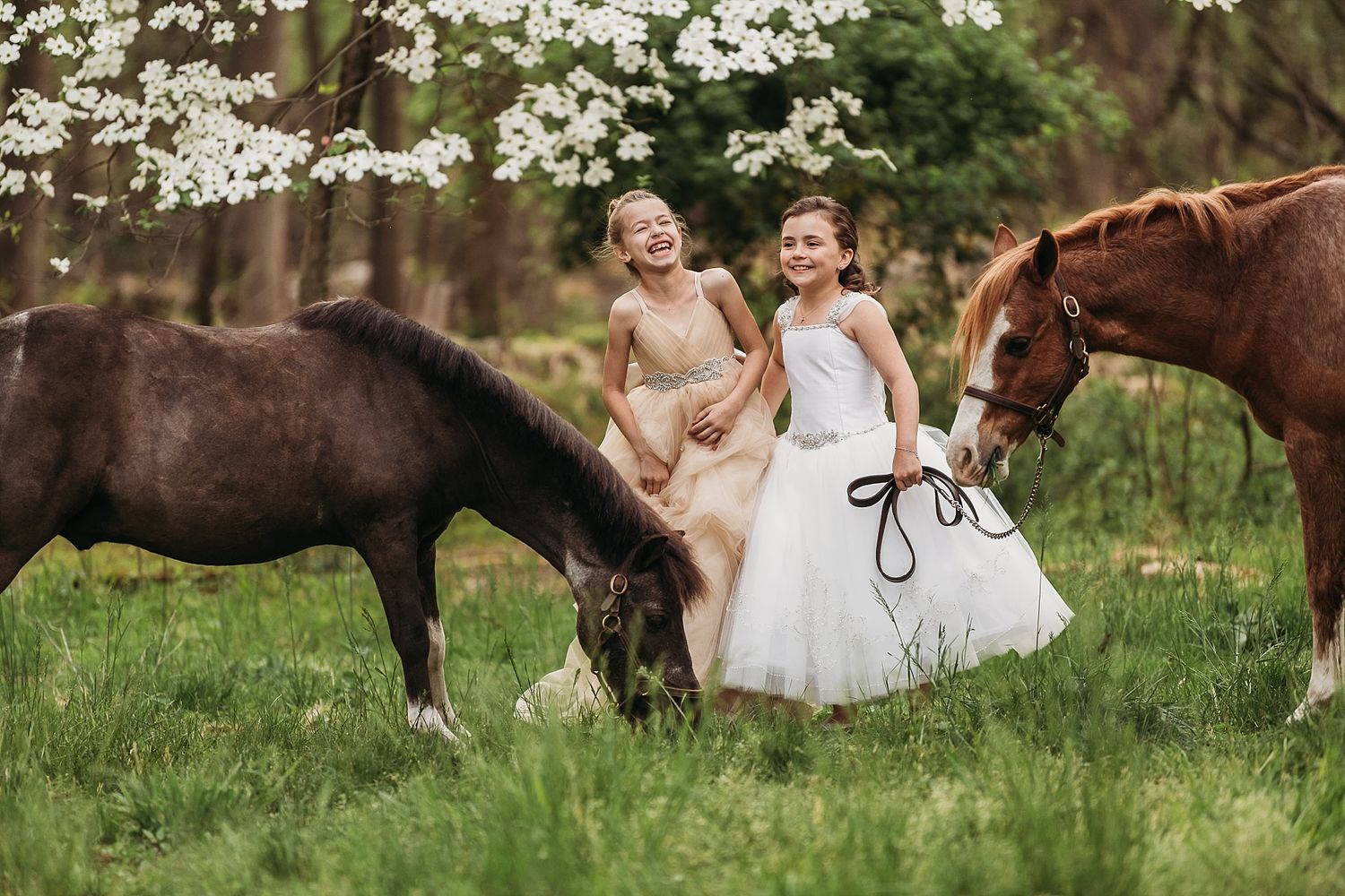Two children in white dresses stand next to grazing horses in a blooming spring field.