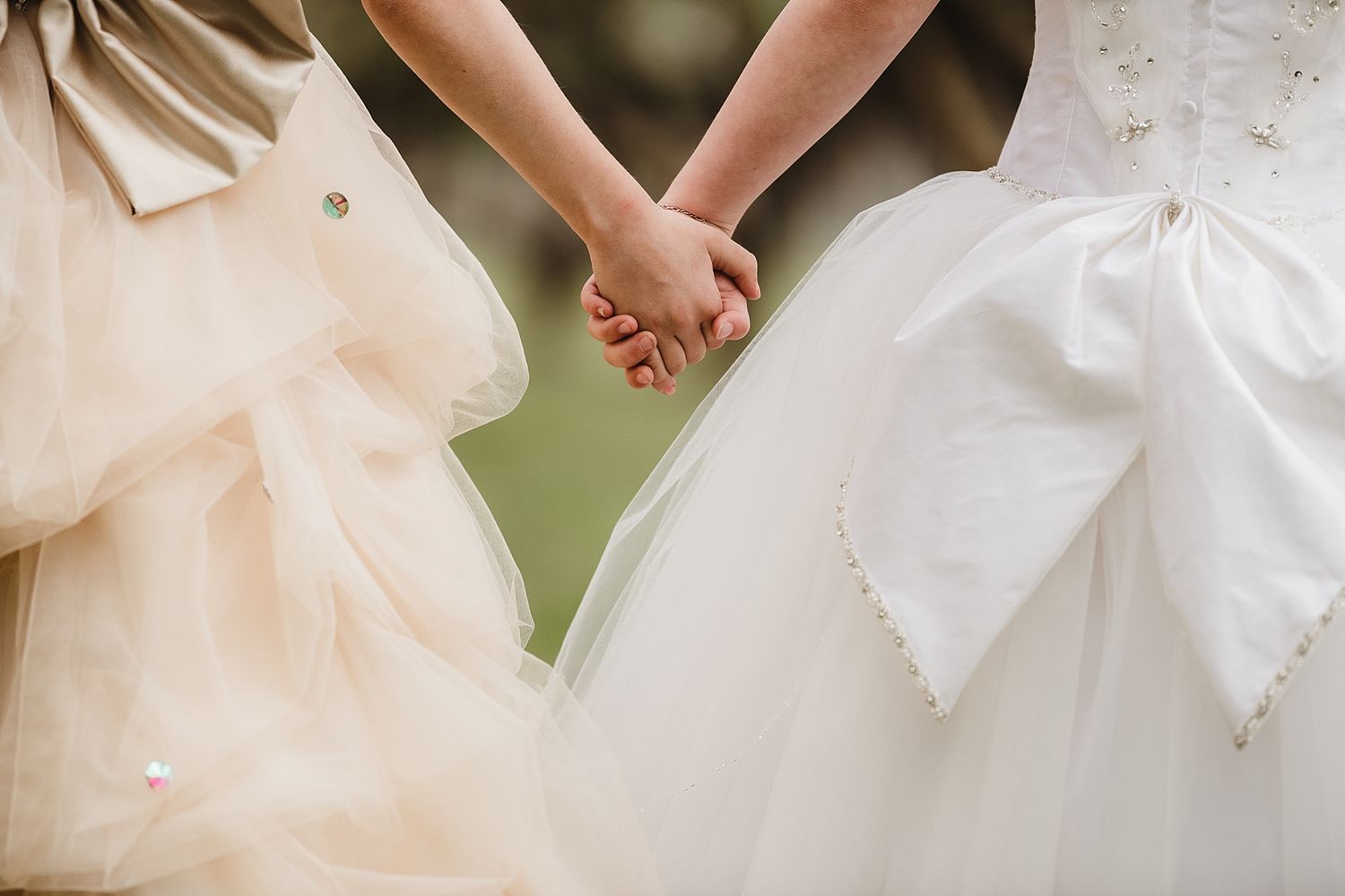 Close-up of two formal dresses side by side with joined hands in soft natural light.