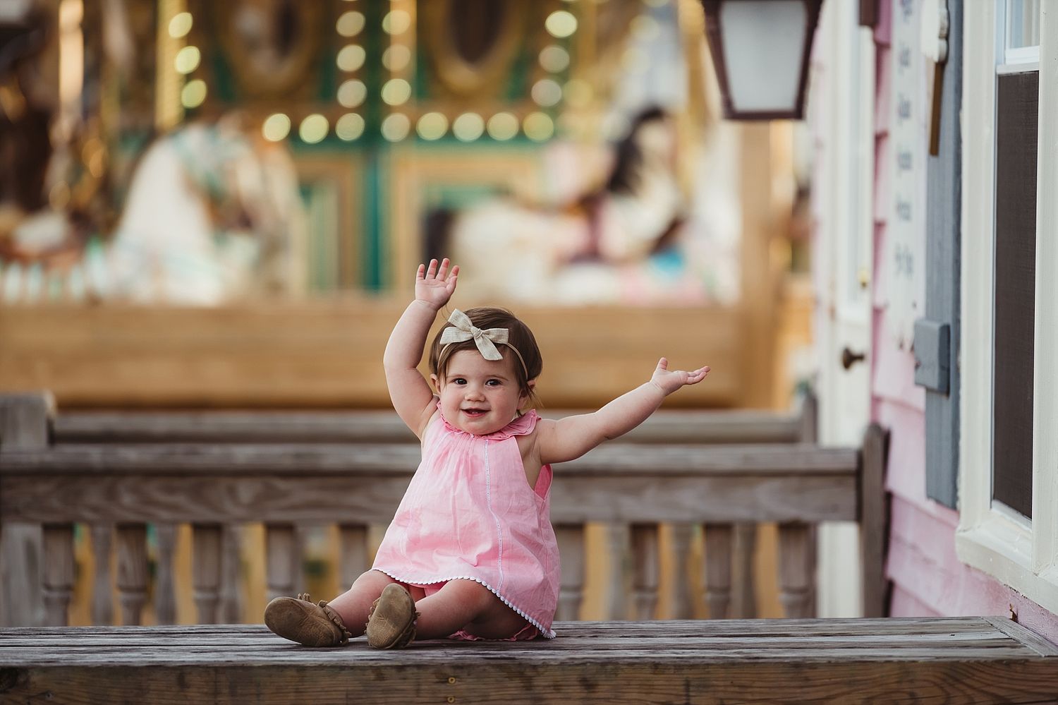 A delighted child in a pink dress spreading arms wide while sitting on wooden bench at a fairground.