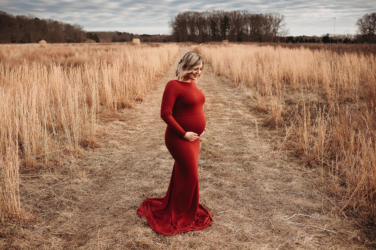 Woman in flowing red dress stands on path through golden autumn field at sunset.