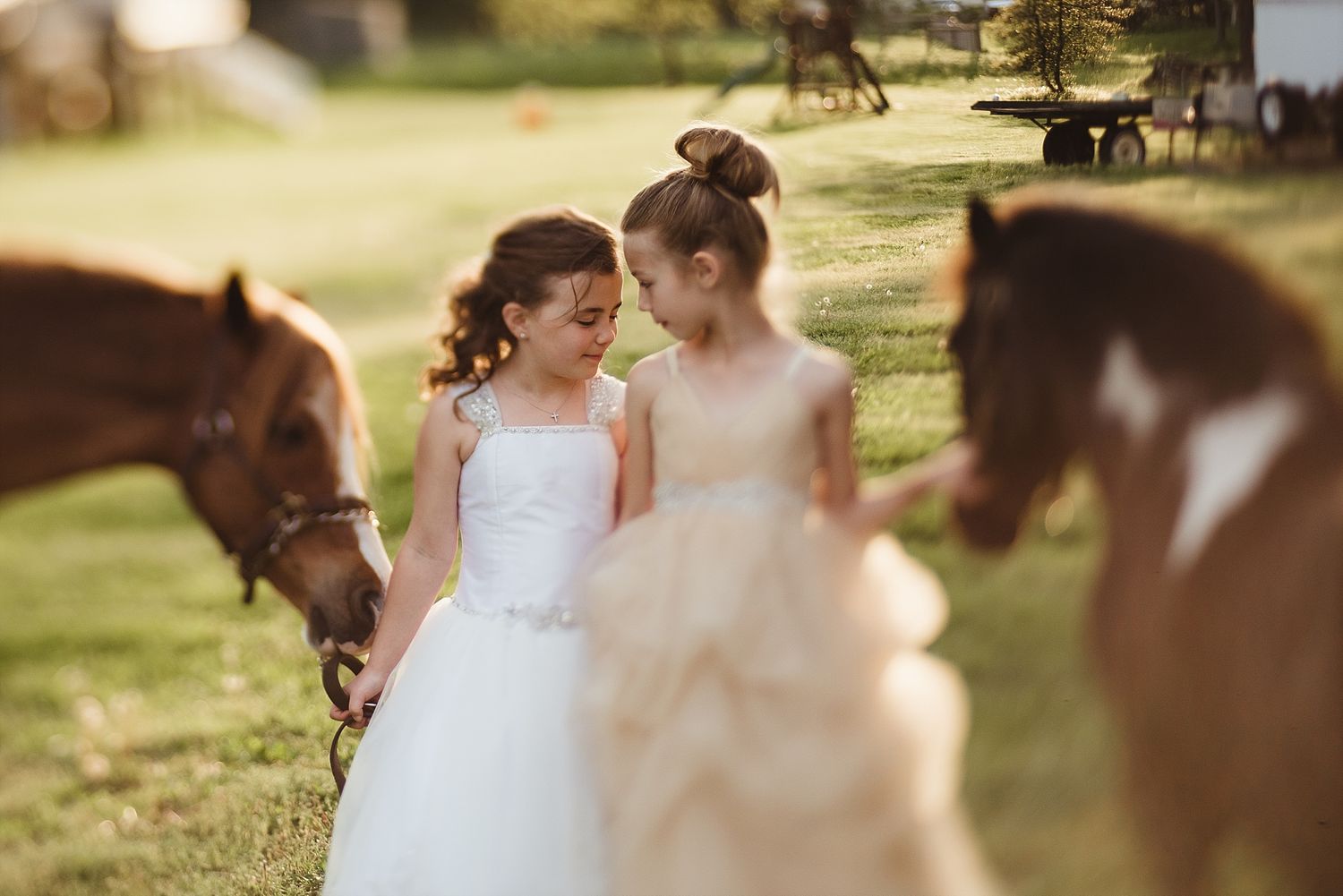 Young girls in white dresses stand near grazing horses in sunlit field.