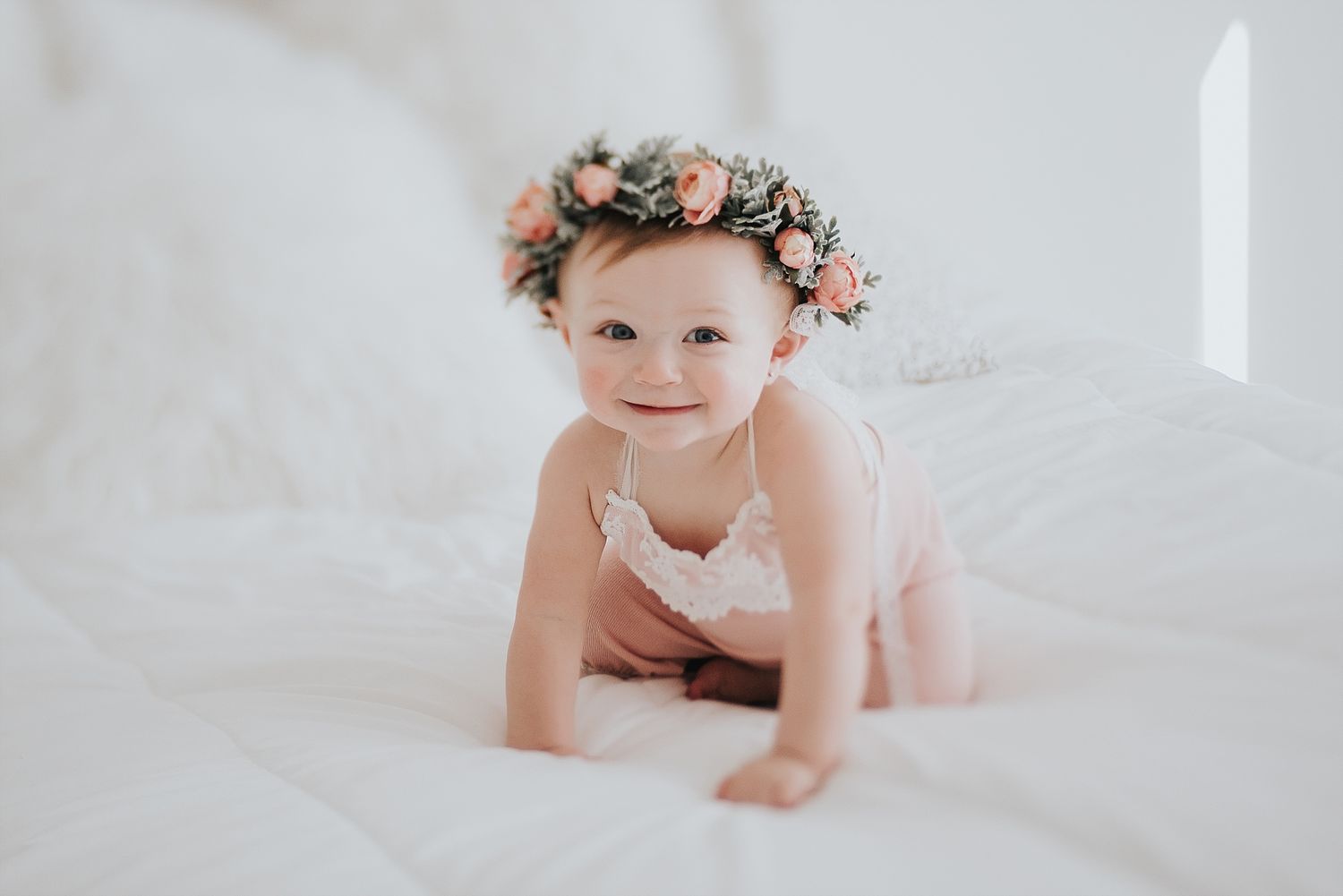 Baby wearing floral crown crawls on white bedding in soft natural light.