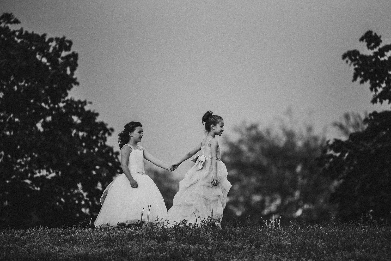 Two figures in white dresses walking together on grassy hillside at dusk.