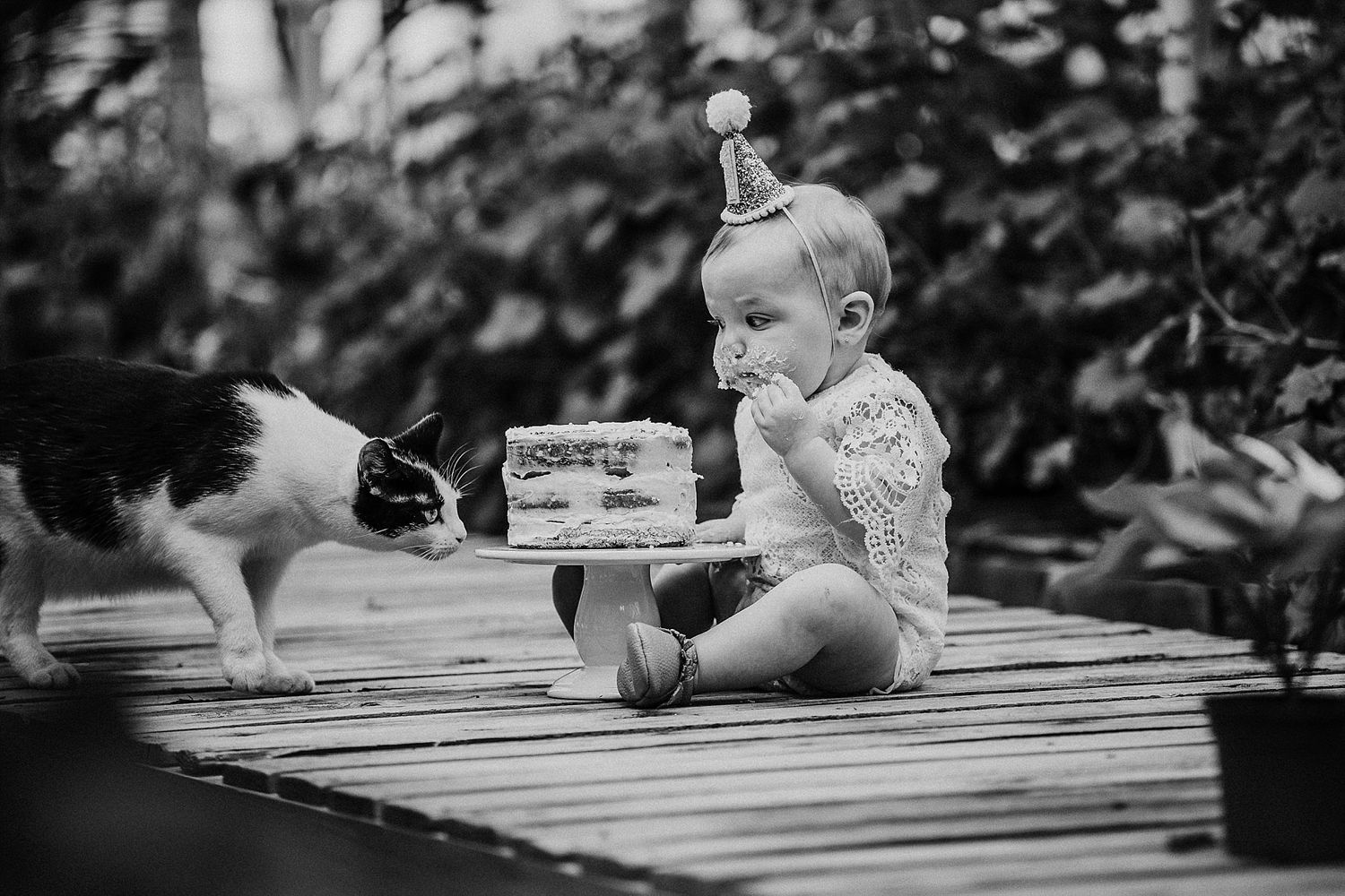 Black and white photo of a baby and cat sitting together on wooden deck with a vintage camera.