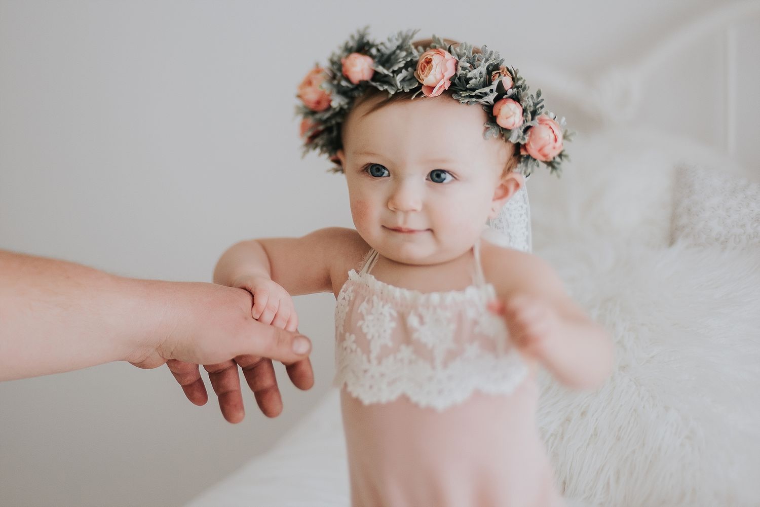 Baby wearing floral crown and lace outfit reaches for outstretched hand.