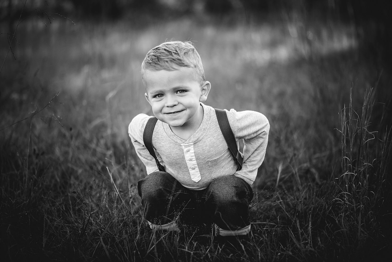 Black and white portrait of young person crouching in tall grass wearing suspenders.