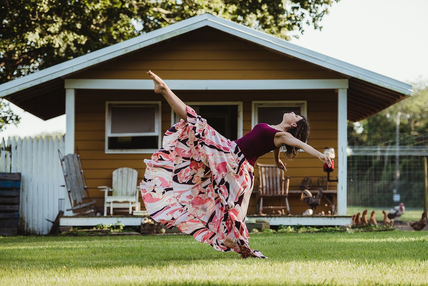 Woman in flowing floral dress dancing gracefully on lawn in front of brown house.