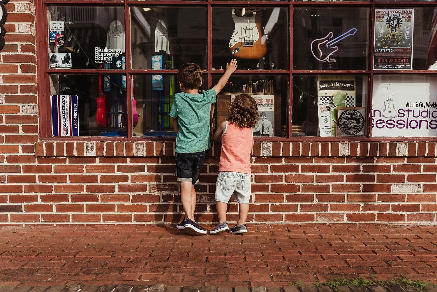 Two children peek through a storefront window display with brick walls and red tiles on the ground.