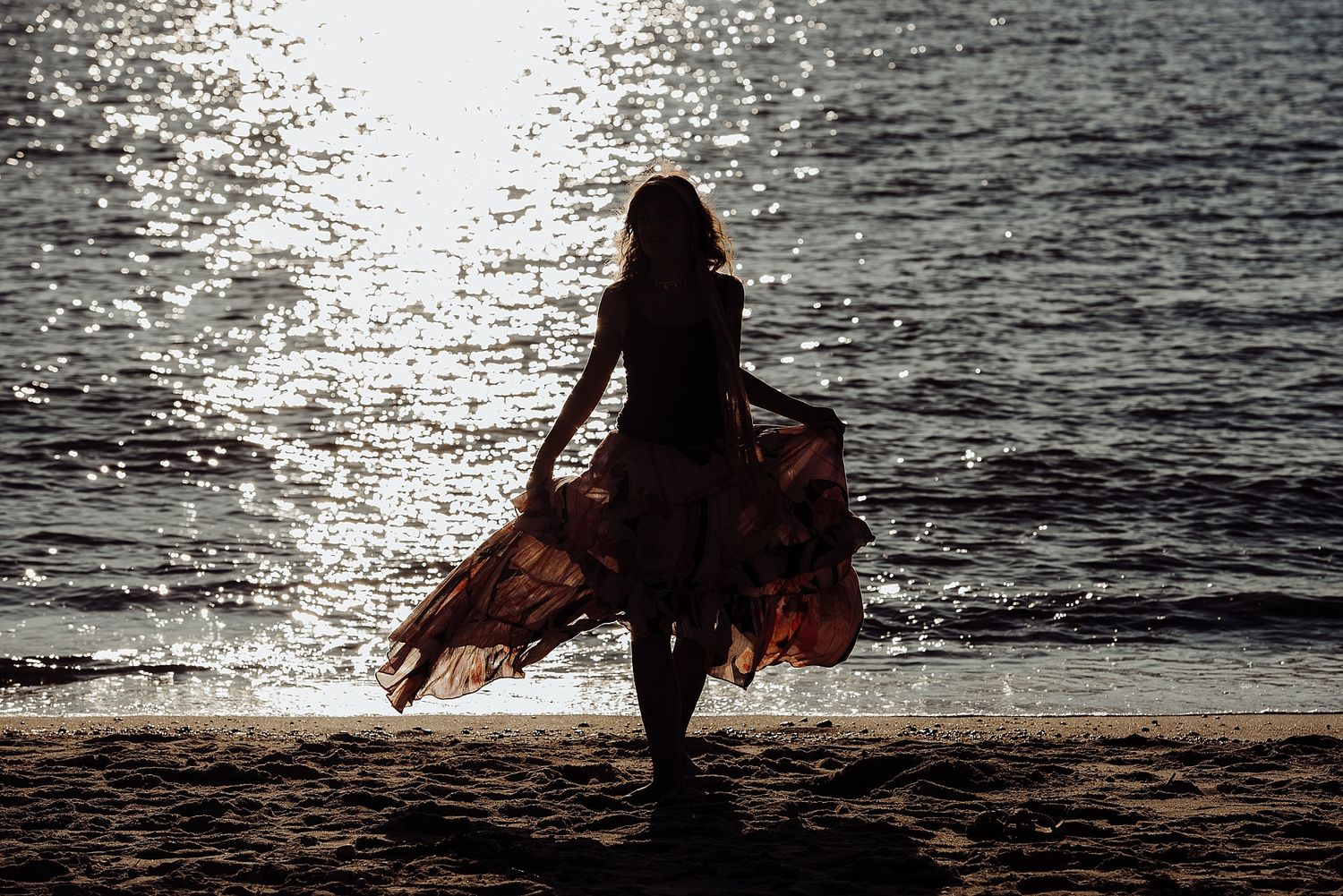 Silhouette of person twirling in flowing dress on beach at sunset with sparkling water.