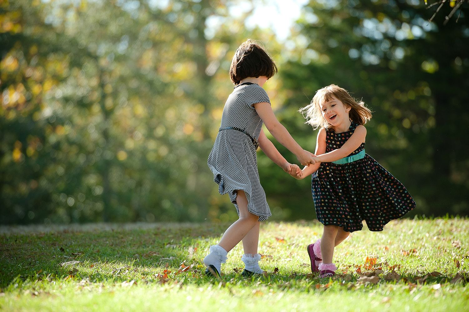 Two children in dresses play together joyfully on a sunny grassy hill.