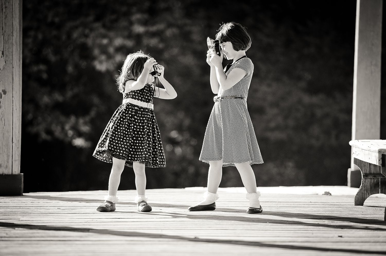Two children in dresses play and pose together in dramatic black and white lighting.