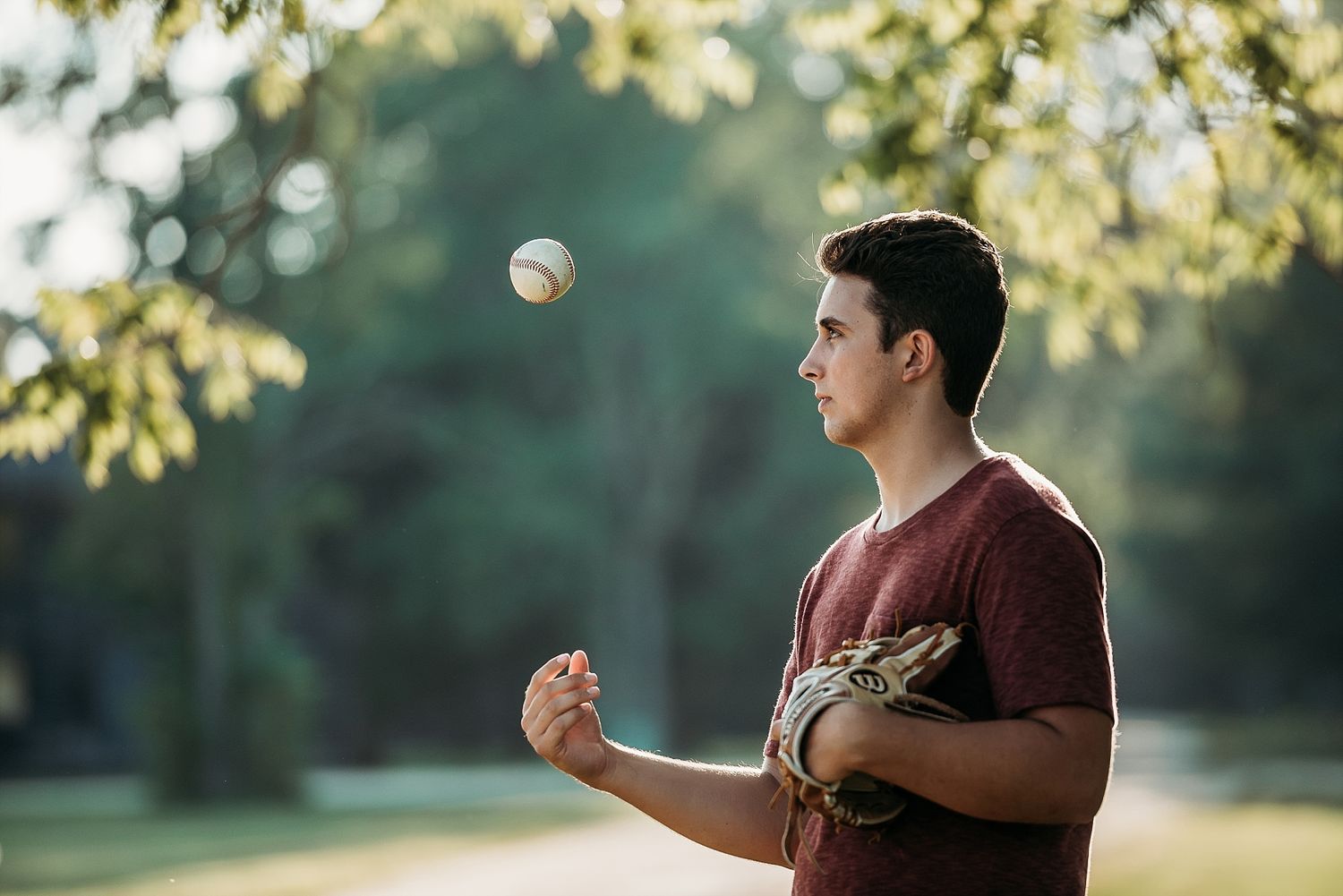 A person in a burgundy shirt practices juggling with a ball in a sunlit park setting.